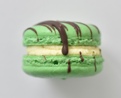Mint chocolate chip French macaron featuring green macaron shells topped with a chocolate drizzle, filled with a mint chocolate buttercream.