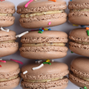 Cosmic Brownie inspired French macaron, featuring brown macaron shells topped with rainbow sprinkles. Filled with a brownie flavored buttercream.