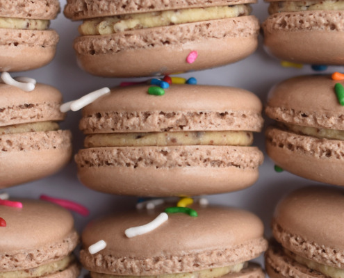 Cosmic Brownie inspired French macaron, featuring brown macaron shells topped with rainbow sprinkles. Filled with a brownie flavored buttercream.